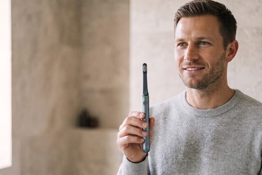 Man smiling and holding a Graphite PulseONE sonic toothbrush in his bathroom