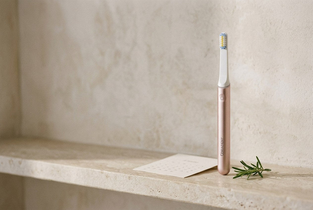 PulseONE sonic toothbrush in Rose standing on a pale stone shelf with a small calendar card and a sprig of rosemary