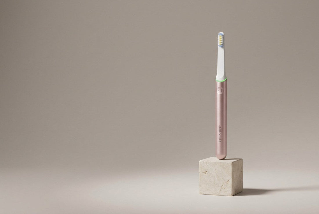 PulseONE sonic toothbrush in Rose standing on a minimal pale stone plinth against a seamless warm grey backdrop