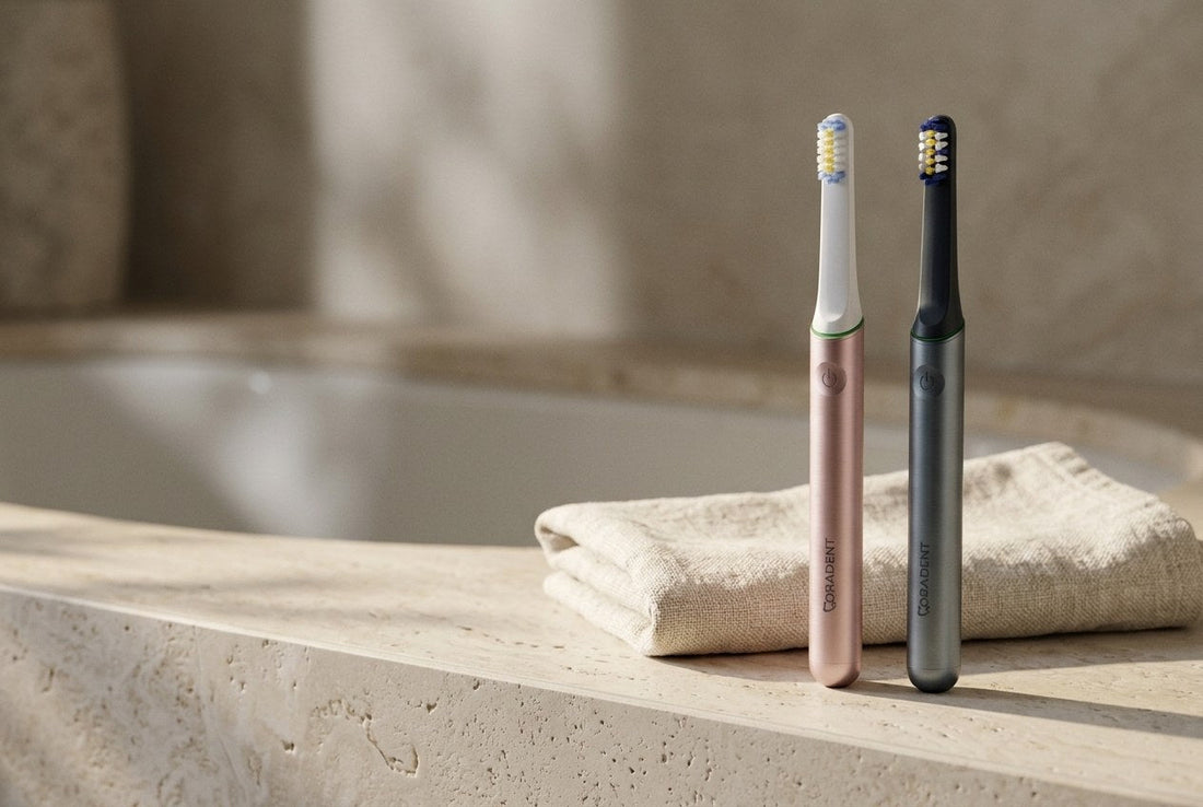 Two PulseONE sonic toothbrushes in Rose and Graphite on a pale travertine bathroom ledge with a folded linen towel