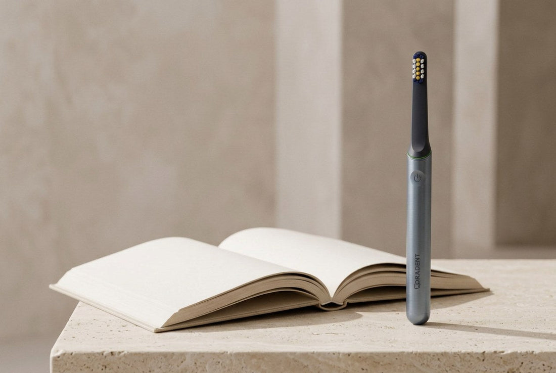 PulseONE sonic toothbrush in Graphite standing on a cream stone surface beside an open book with blank pages