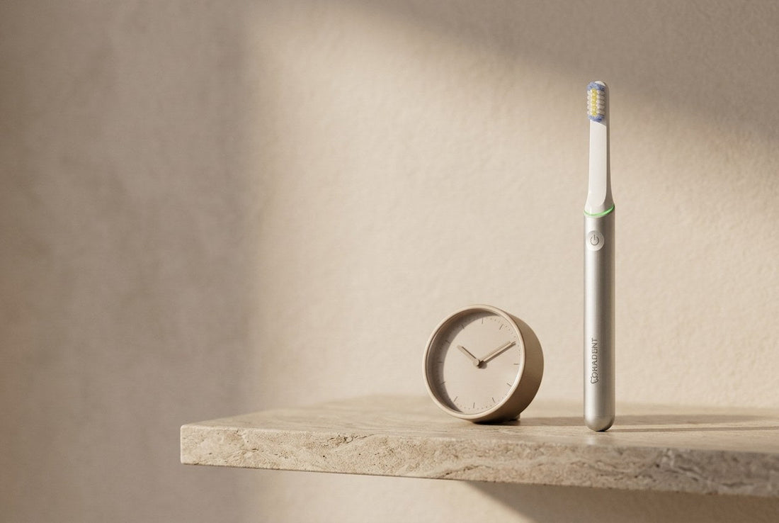 PulseONE sonic toothbrush in Platinum standing beside a minimalist round analogue clock lying on a stone shelf