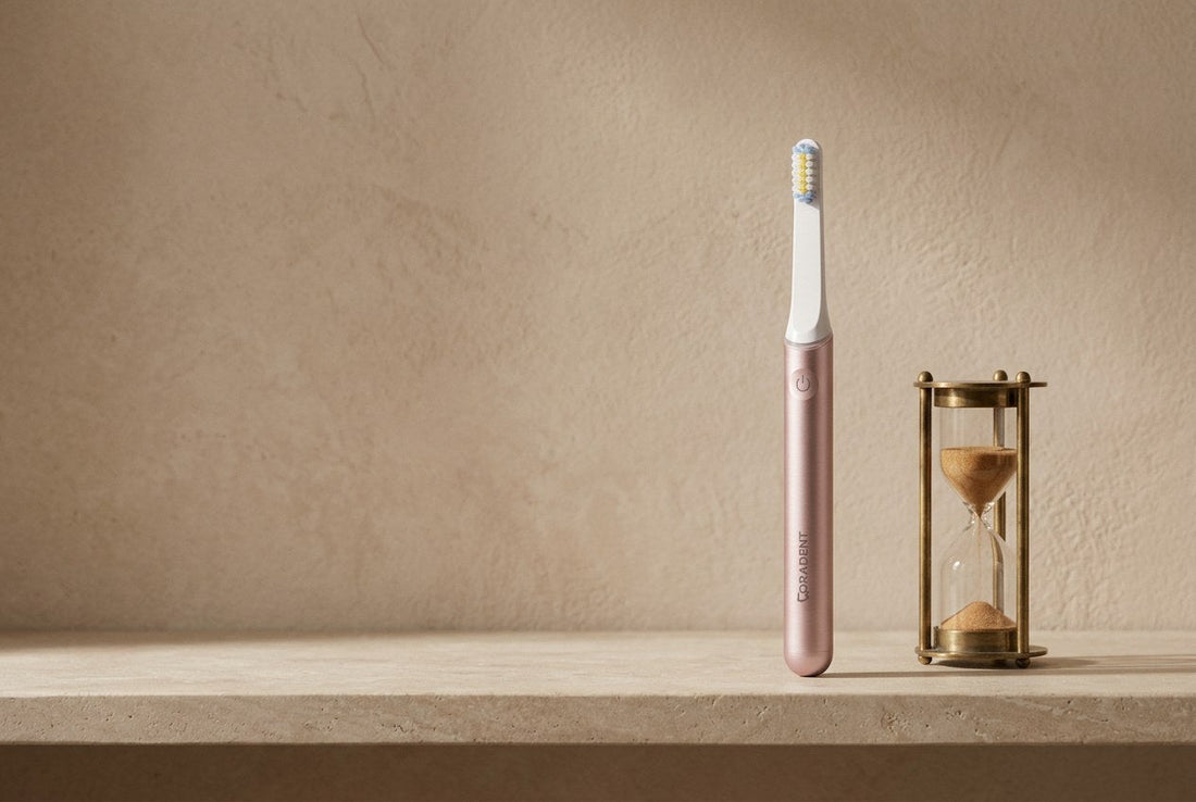 PulseONE sonic toothbrush in Rose standing on a honed limestone shelf next to a small brass sand timer