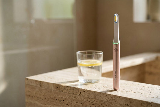 PulseONE sonic toothbrush in Rose standing on a travertine ledge beside a glass of water with a slice of lemon